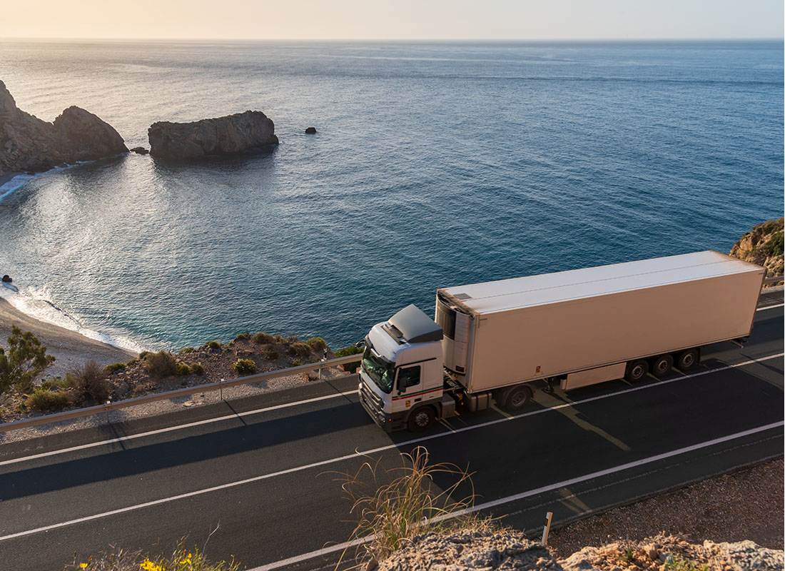 Insurance by Industry - View of a White Truck with a Trailer Driving Down a Road Up on a Cliff with a Scenic View of the Beach Below