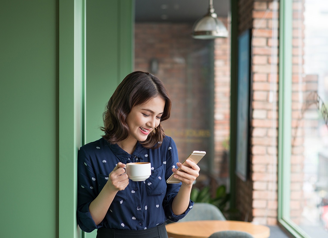 Read Our Reviews - Portrait of a Young Smiling Woman Using her Phone While Holding a Cup of Coffee Inside a Small Cafe